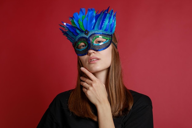 Linda jovencita con máscara de carnaval azul y soporte sobre fondo rojo