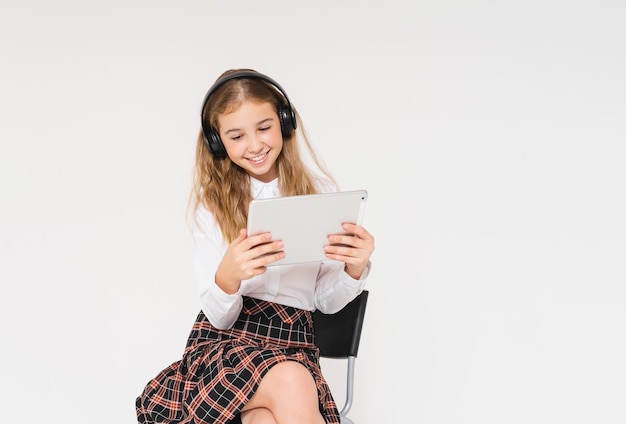 Linda jovencita sonriente en uniforme escolar con auriculares y una tableta en sus manos