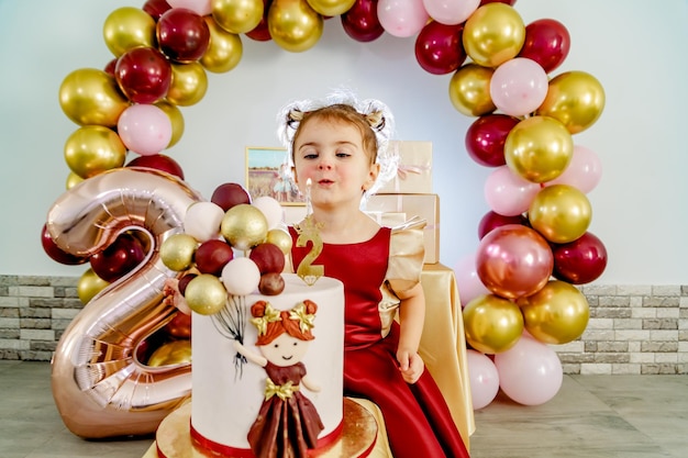 Una linda y linda niña de dos años con un vestido rojo apaga una vela en un pastel de cumpleaños