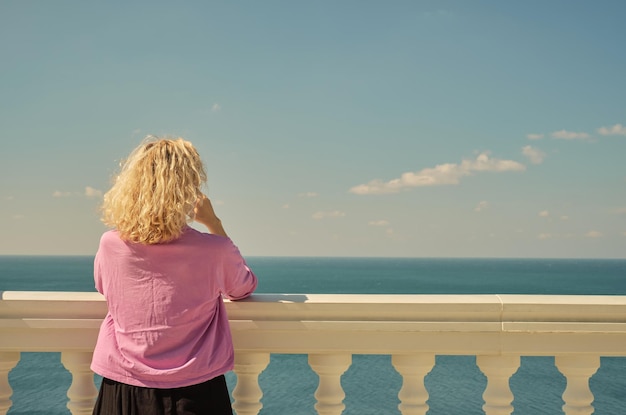 Linda loira adulta em uma camiseta rosa fica e desfruta de uma vista para o mar da varanda espaço de visão traseira para texto