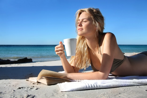 Linda loira bebendo uma xícara de café e lendo um livro na praia