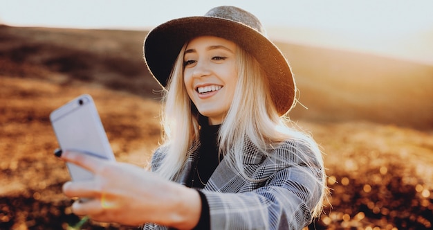 Linda loira caucasiana com lindo chapéu fazendo uma selfie enquanto sorri em um campo ensolarado