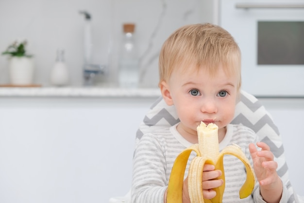 Linda loira caucasiana infantil cerca de um ano sentado na cadeira, almoçando na cozinhabebê e ...