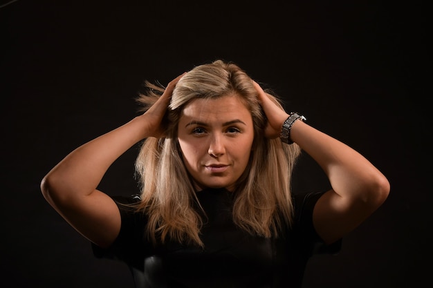 Linda loira com cabelo comprido olha para a câmera e penteia o cabelo com as mãos em um fundo preto. Fotografado de frente