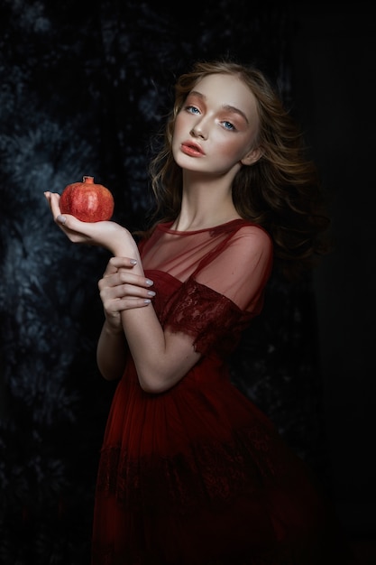 Linda loira com fruta romã nas mãos dela. Retrato de primavera de uma menina em um vestido vermelho, quebrando uma romã, suco fluindo pelas mãos dela