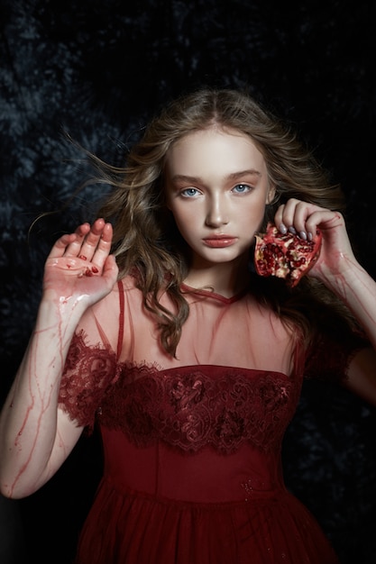 Linda loira com fruta romã nas mãos dela. Retrato de primavera de uma menina em um vestido vermelho, quebrando uma romã, suco fluindo pelas mãos dela