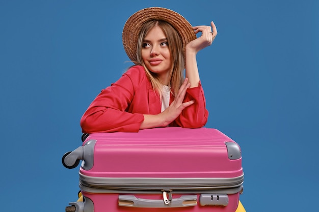 Linda loira de chapéu de palha blusa branca jaqueta vermelha Ela está sorrindo enquanto está sentado encostado em malas coloridas posando em fundo azul Conceito de viagem Closeup cópia espaço