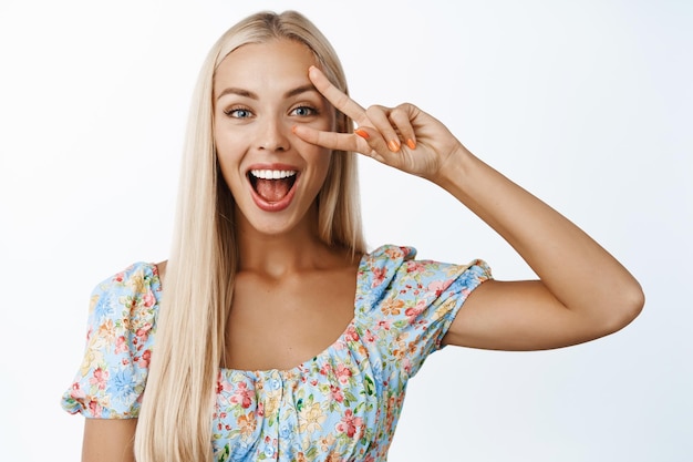 Linda loira entusiástica mostrando paz vsign sorrindo e parecendo espantada em pé sobre fundo branco