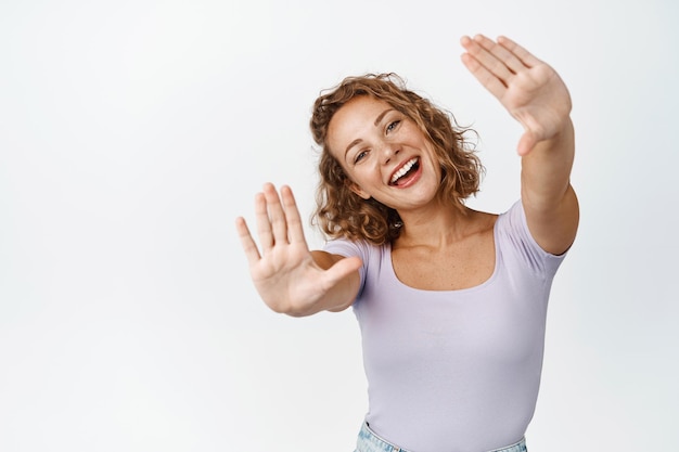 Linda loira imaginando algo, esticando as mãos em gesto livre e feliz, sorrindo e rindo, de pé sobre fundo branco.