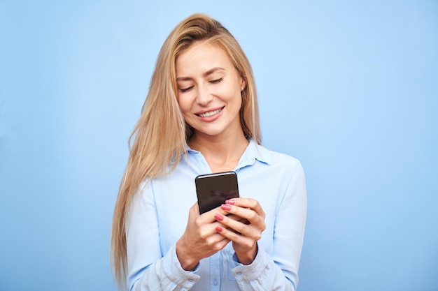 Linda loira olhando para a tela do smartphone e sorrindo isolada no fundo azul do estúdio