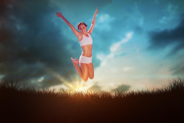 Linda loira pulando com os braços abertos contra o céu azul sobre a grama