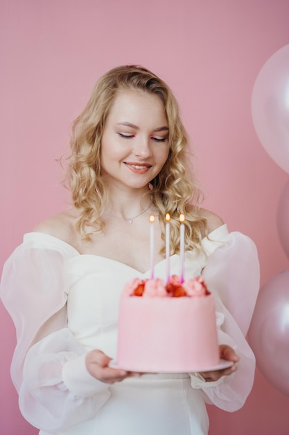 linda loira segurando um bolo de aniversário em um fundo rosa, as velas no bolo
