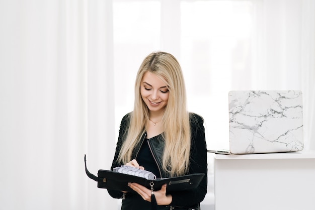 Linda loira sorridente com um caderno e caneta nas mãos dela, ao lado de um laptop em uma janela de luz, empregado de escritório