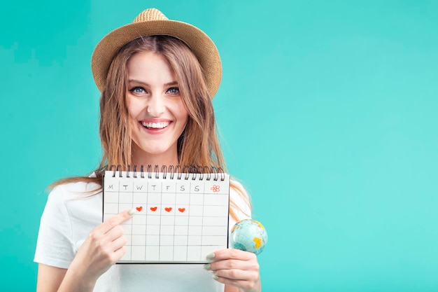 Linda loira usa camiseta branca e chapéu segurando o calendário de férias no conceito de destino de verão de fundo azul