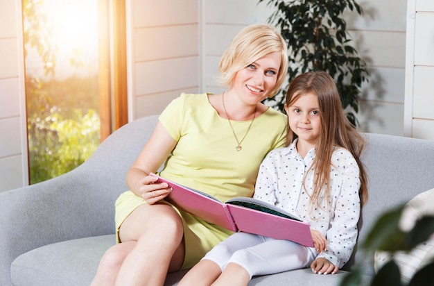 Linda madre e hija sudada sentadas en el sofá de la casa moderna y leyendo un libro