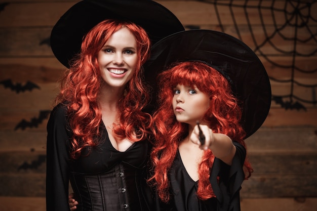 Linda mãe caucasiana e sua filha com longos cabelos vermelhos em trajes de bruxa e varinha mágica comemorando Halloween posando
