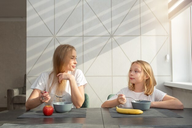 linda mãe com filha na cozinha em casa na mesa comendo mingau