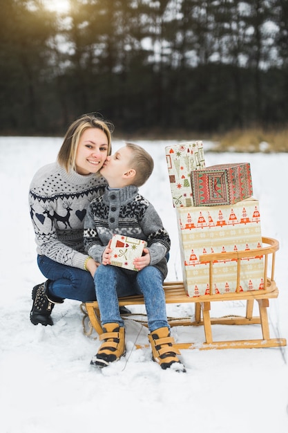 Linda mãe com o filho durante um passeio de trenó em winter park, sentada em um trenó de madeira com presentes