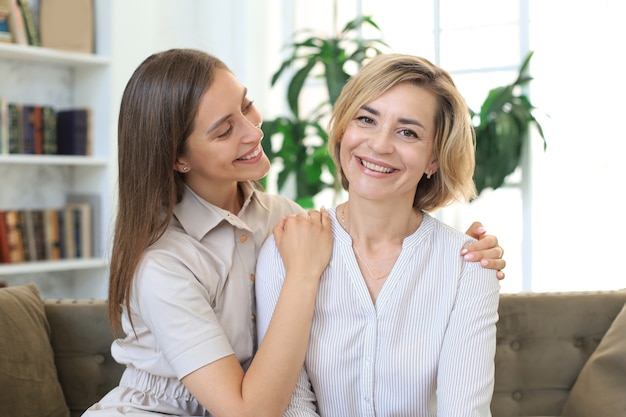 Linda mãe de meia-idade e sua filha adulta estão se abraçando e sorrindo enquanto está sentado no sofá em casa.