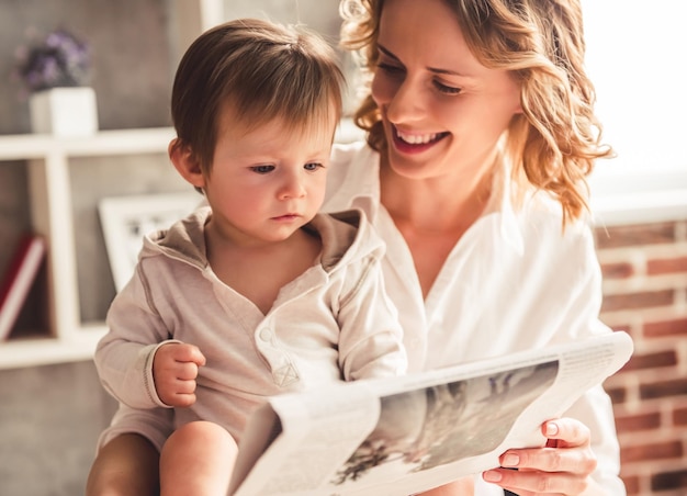 Linda mãe de negócios de terno está lendo um jornal com seu bebê fofo e sorrindo em casa