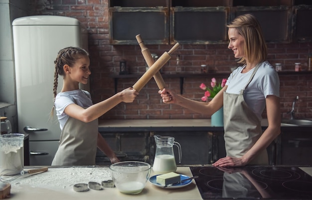 Linda mãe e filha em aventais estão fingindo lutar com rolos olhando para a câmera e sorrindo enquanto cozinha em casa