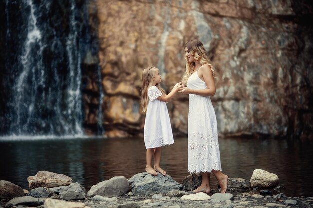 Linda mãe e filha vestindo vestidos brancos nas montanhas perto do rio no fundo de uma cachoeira