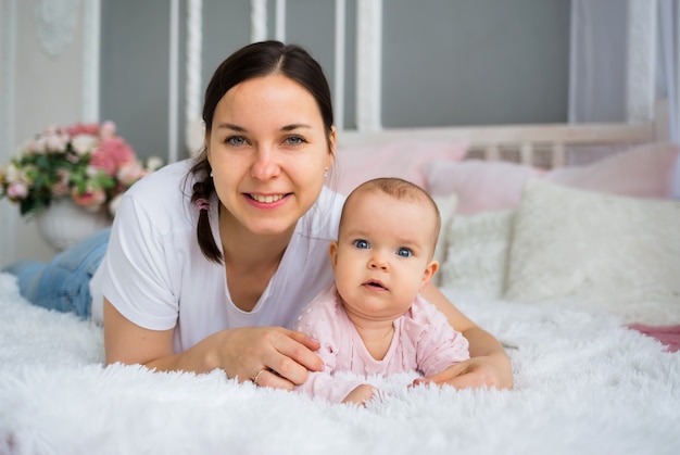 Linda mãe morena com uma camiseta branca deita-se na cama e abraça sua filha bebê com um vestido rosa