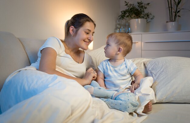 Linda mãe sorridente deitada na cama e brincando com seu bebê antes de dormir