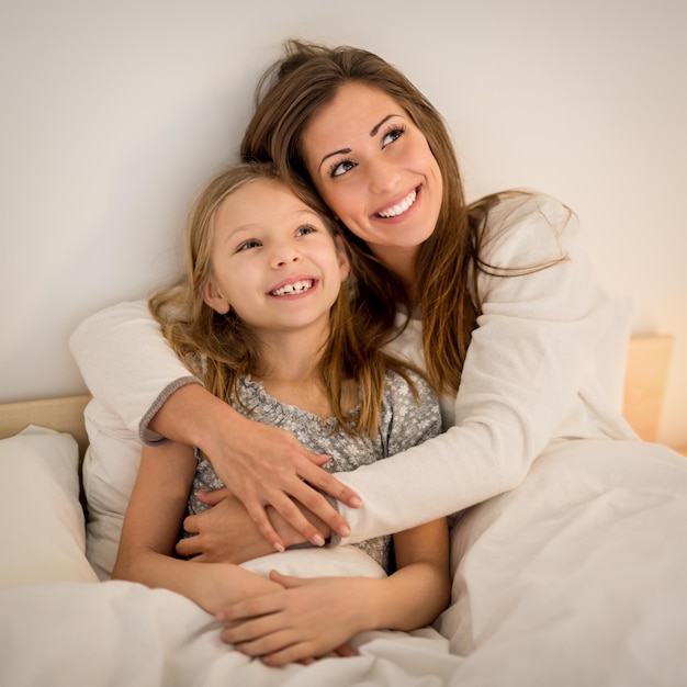 Linda mãe sorridente e sua filha abraçando na cama. Eles estão sentados na cama de pijama e sorrindo.