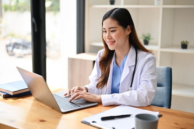 Linda médica asiática de uniforme usando seu laptop em seu espaço de trabalho em um escritório de hospital