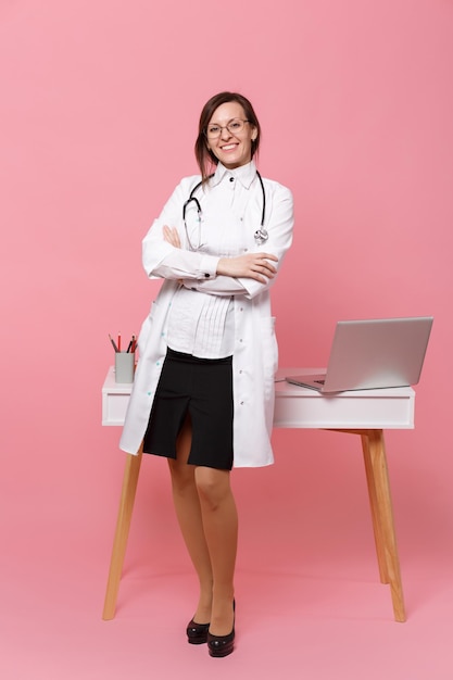 Linda médica fica na frente da mesa com o computador pc, documento médico no hospital isolado no fundo da parede rosa pastel. Mulher de estetoscópio de óculos de vestido médico. Conceito de medicina de saúde