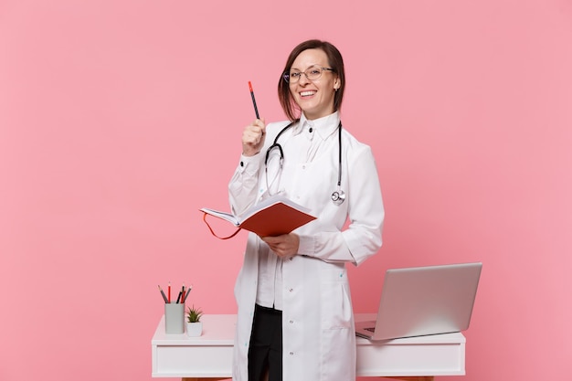 Linda médica fica na frente da mesa com o computador pc, documento médico no hospital isolado no fundo da parede rosa pastel. Mulher de estetoscópio de óculos de vestido médico. Conceito de medicina de saúde