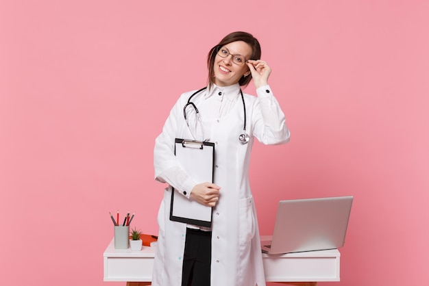 Linda médica fica na frente da mesa com o computador pc, documento médico no hospital isolado no fundo da parede rosa pastel. Mulher de estetoscópio de óculos de vestido médico. Conceito de medicina de saúde