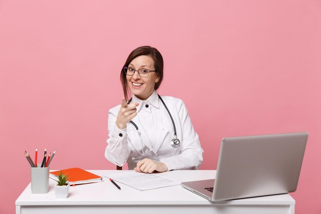 Linda médica se senta na mesa trabalha no computador com o documento médico no hospital isolado no fundo da parede rosa pastel. mulher de estetoscópio de óculos de vestido médico. conceito de medicina de saúde