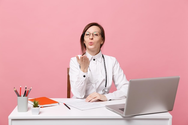 Linda médica se senta na mesa trabalha no computador com o documento médico no hospital isolado no fundo da parede rosa pastel. Mulher de estetoscópio de óculos de vestido médico. Conceito de medicina de saúde