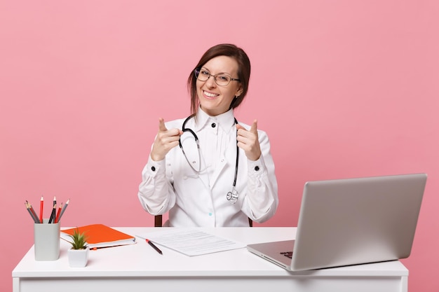 Linda médica se senta na mesa trabalha no computador com o documento médico no hospital isolado no fundo da parede rosa pastel. Mulher de estetoscópio de óculos de vestido médico. Conceito de medicina de saúde