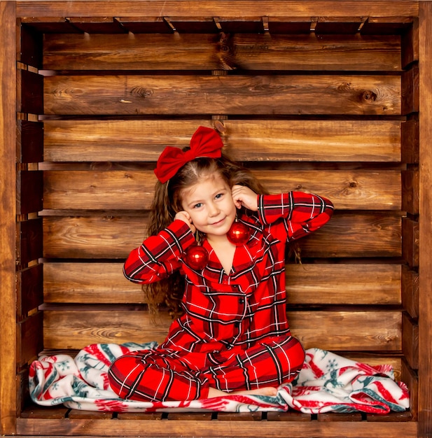 Linda menina alegre com pijama de natal xadrez vermelho segurando bolas de natal em um fundo de madeira