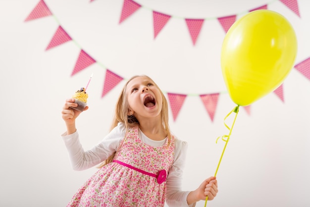 Linda menina alegre segurando balão e bolo de aniversário pequeno com vela.