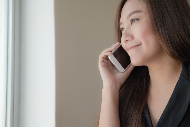 Linda menina asiática falando no telefone inteligente com carinha no escritório