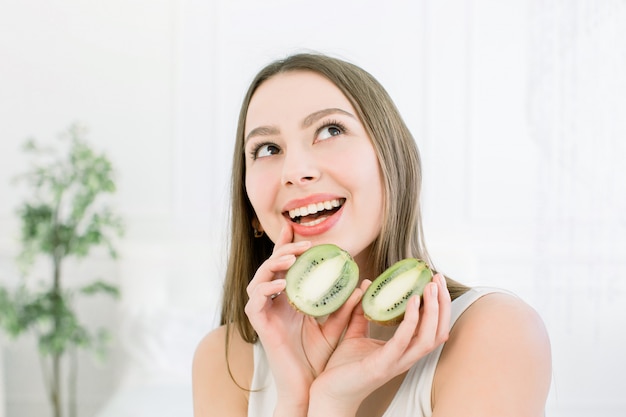 Linda menina bonita, segurando uma fatia de kiwi perto da cabeça e sorrindo. Alimentos saudáveis para a pele fresca. Conceito de beleza