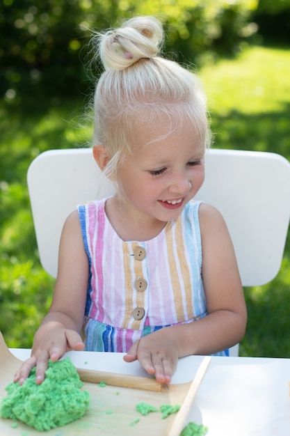 Linda menina com cabelo loiro brincando com areia cinética colorida ao ar livre no quintal em dia de verão. Conceito de passatempo, atividade de lazer, artesanato, modelagem e passatempo criativo