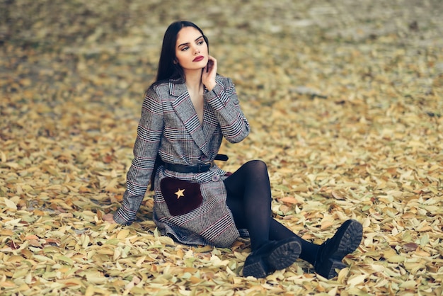 Linda menina com cabelo muito comprido, vestindo casaco de inverno, sentado no chão de um parque urbano