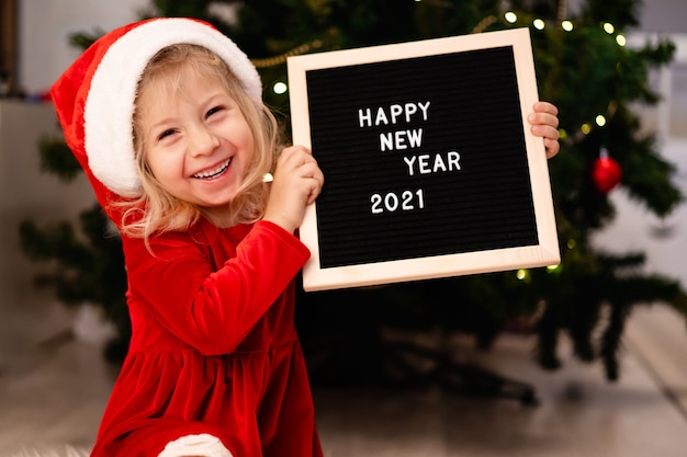 Foto linda menina com chapéu de papai noel e vestido de natal vermelho está deitada sob a árvore de natal e rindo, ao lado de um quadro preto e um texto com letras brancas feliz ano novo 2021. foto de alta qualidade