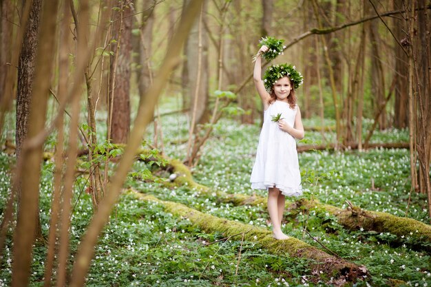 Linda menina com um vestido branco caminha na floresta de primavera. retrato da menina bonita com uma coroa de flores da primavera na cabeça.