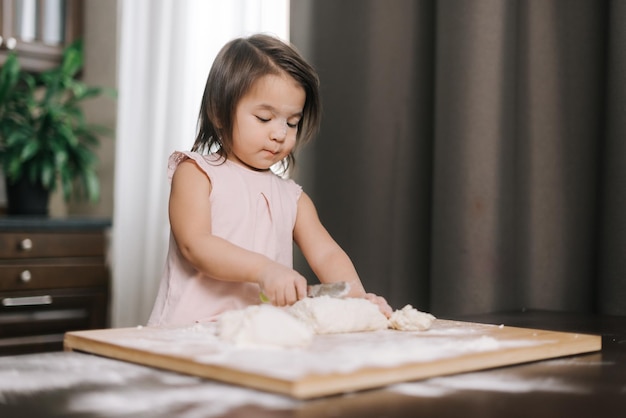Linda menina curiosa está cortando massa com faca de criança na mesa na cozinha com interior moderno. Criança prepara massa para assar biscoitos