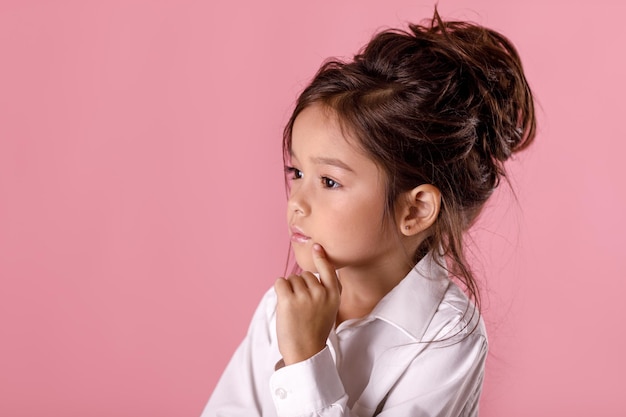 Linda menina duvidosa e pensativa em uma camisa branca com penteado em fundo rosa. criança pensando. emoções humanas