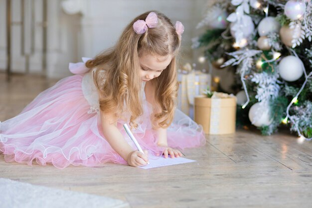 Linda menina escrevendo carta para o Papai Noel na véspera de ano novo. criança bonita está pensando debaixo da árvore de Natal.