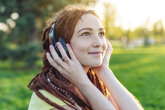 Linda menina moderna com dreadlocks ouvindo música com fones de ouvido no outono sunny park