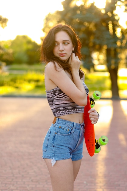 Linda menina morena ativa com cabelo comprido, vestindo top, shorts jeans azul e tênis rosa elegantes, posando no fundo do sol. segurando um skate laranja com rodas verdes.