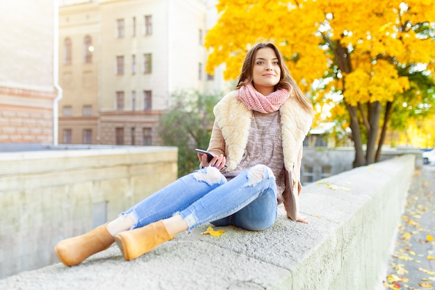 Linda menina morena caucasiana, sentado dia quente de outono com fundo de árvores com folhagem amarela e uma cidade